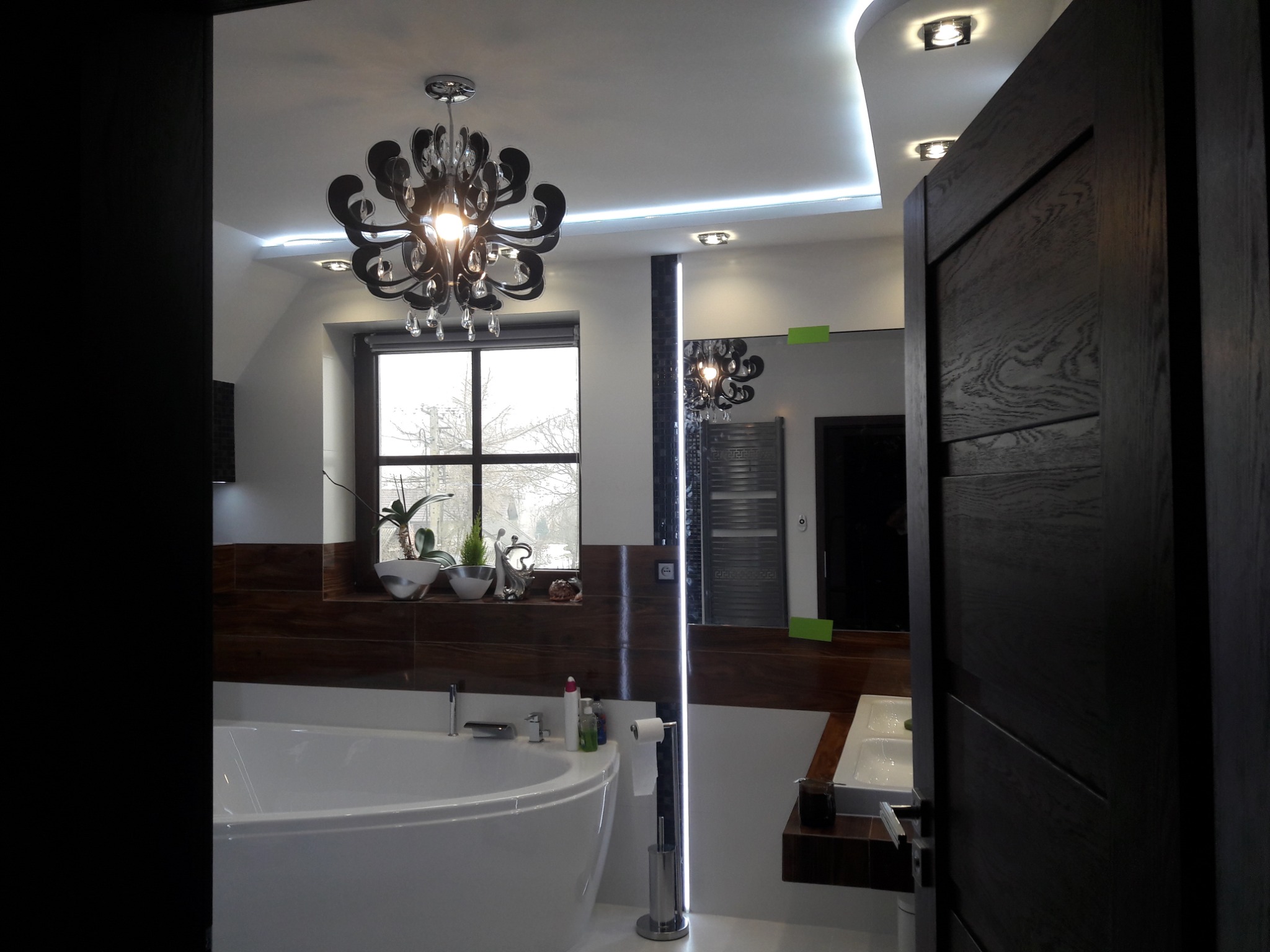 Nowoczesna łazienka z owalną wanną, ciemnymi drewnianymi panelami, dużym lustrem i ozdobnym żyrandolem w kształcie kwiatu, oświetlona światłem LED