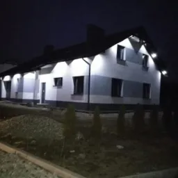 Oświetlona elewacja dwukondygnacyjnego domu w nocy, z widocznym fragmentem ogrodu i chodnika.