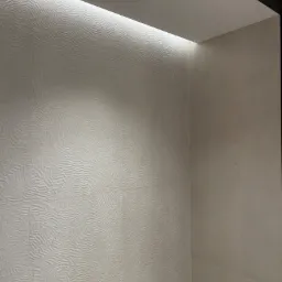 Ściana łazienki wykończona dekoracyjnymi, trójwymiarowymi płytami w odcieniach bieli, o nieregularnej fakturze przypominającej piaskowe wydmy, oświetlona listwą LED ukrytą w suficie.