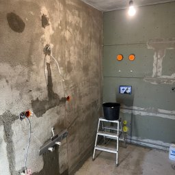 „JUPINEX “ Adam Jueppner - Surowa łazienka w trakcie prac: ściany z szarym tynkiem i płytami gips-karton, widoczne instalacje elektryczne i wodne, drabina z wiadrem. Oświetlenie żarówką.