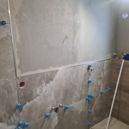 „JUPINEX “ Adam Jueppner - Układanie płytek w łazience z użyciem systemu poziomowania. Widoczne płytki imitujące marmur, listwa dekoracyjna i przygotowane punkty instalacyjne.