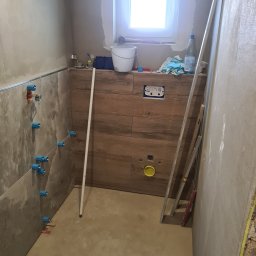 „JUPINEX “ Adam Jueppner - Łazienka w trakcie remontu: częściowo ułożone płytki imitujące drewno, widoczne instalacje, poziomica oparta o ścianę. Prace wykończeniowe w toku.