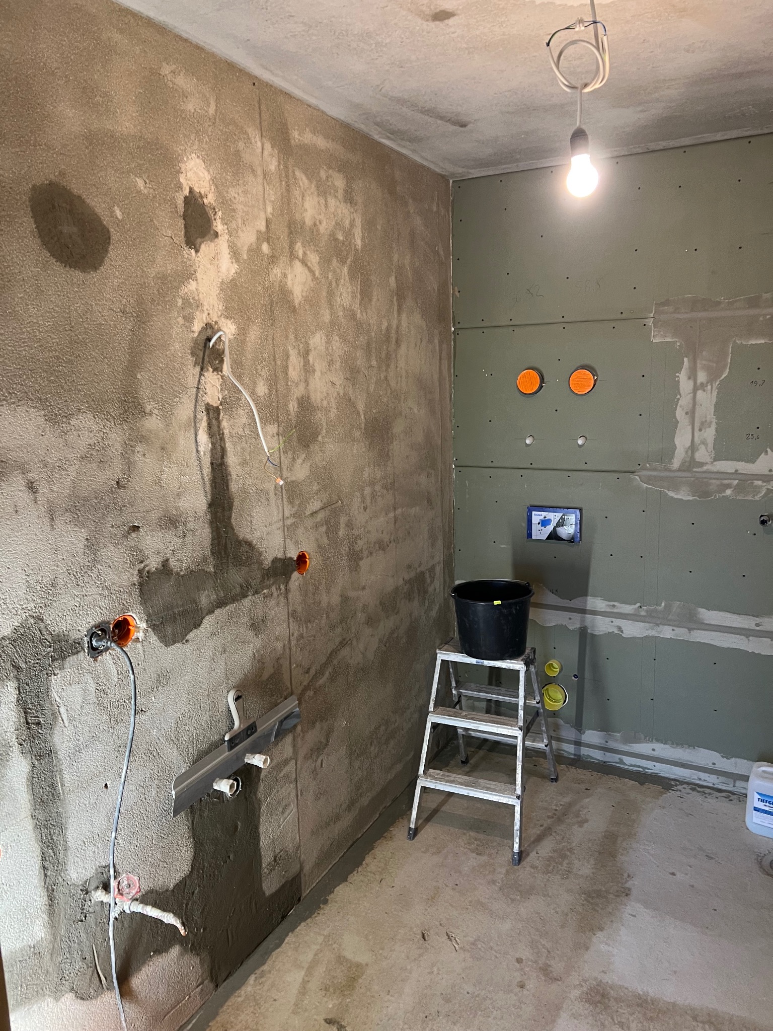 Surowa łazienka w trakcie prac: ściany z szarym tynkiem i płytami gips-karton, widoczne instalacje elektryczne i wodne, drabina z wiadrem. Oświetlenie żarówką.