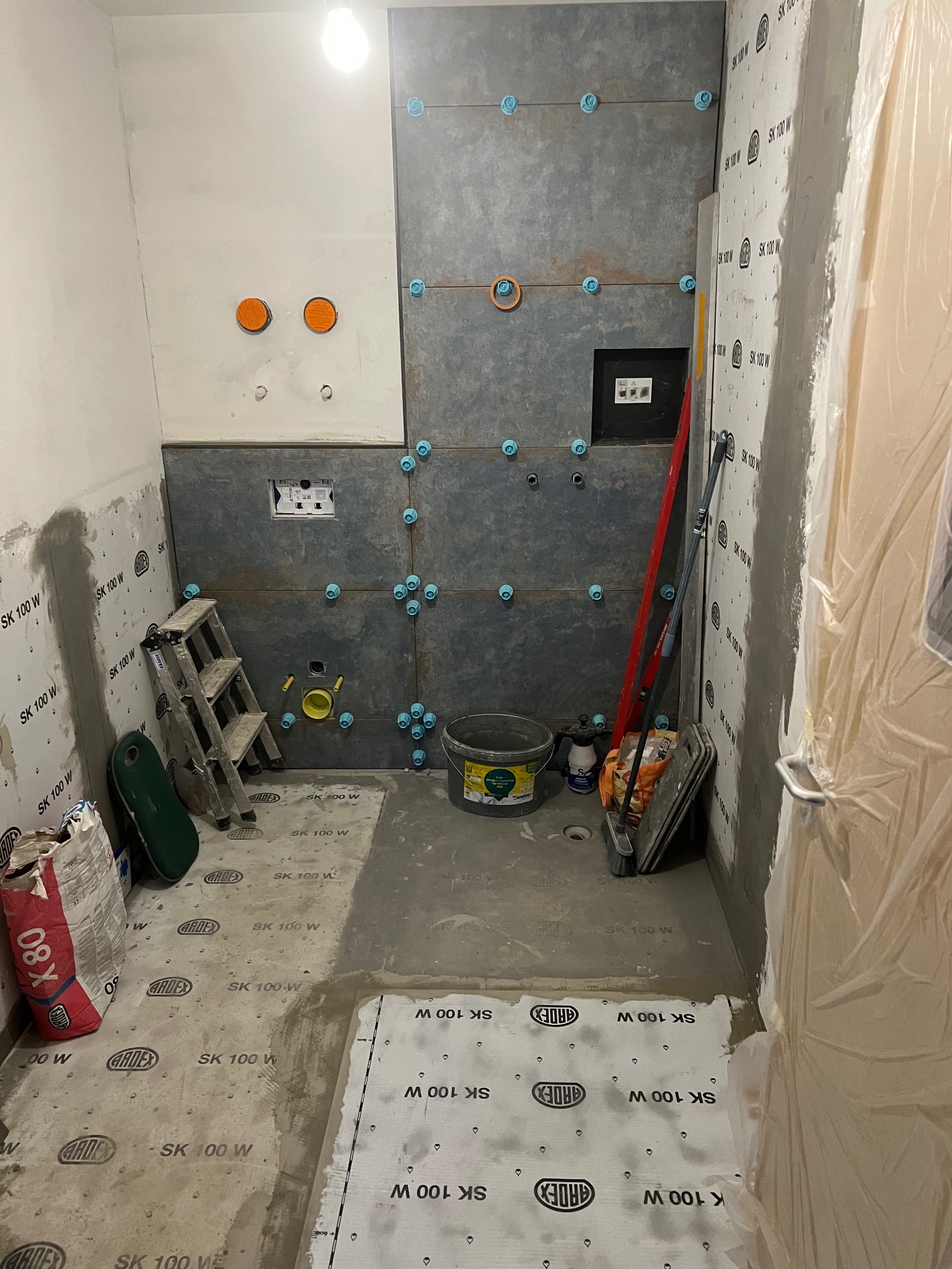 Łazienka w trakcie remontu: szare płytki z niebieskimi krzyżykami dystansowymi na ścianie, widoczne narzędzia, worki z zaprawą i folia ochronna na drzwiach.