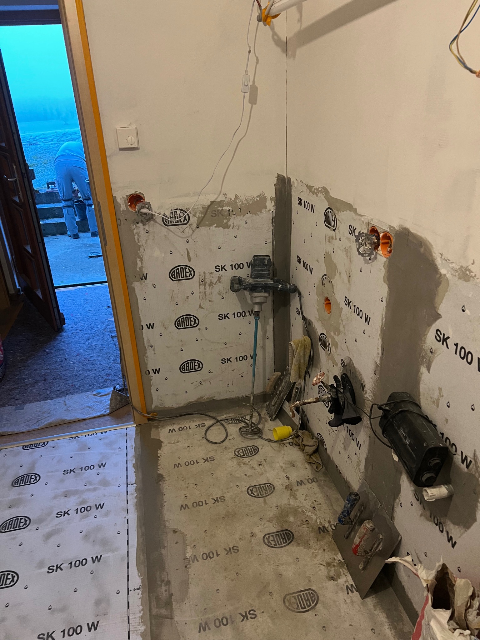 Przygotowanie ścian pod płytki w łazience: hydroizolacja folią w płynie Ardex SK 100 W, widoczne puszki elektryczne, mieszadło i narzędzia glazurnicze. Praca w toku.
