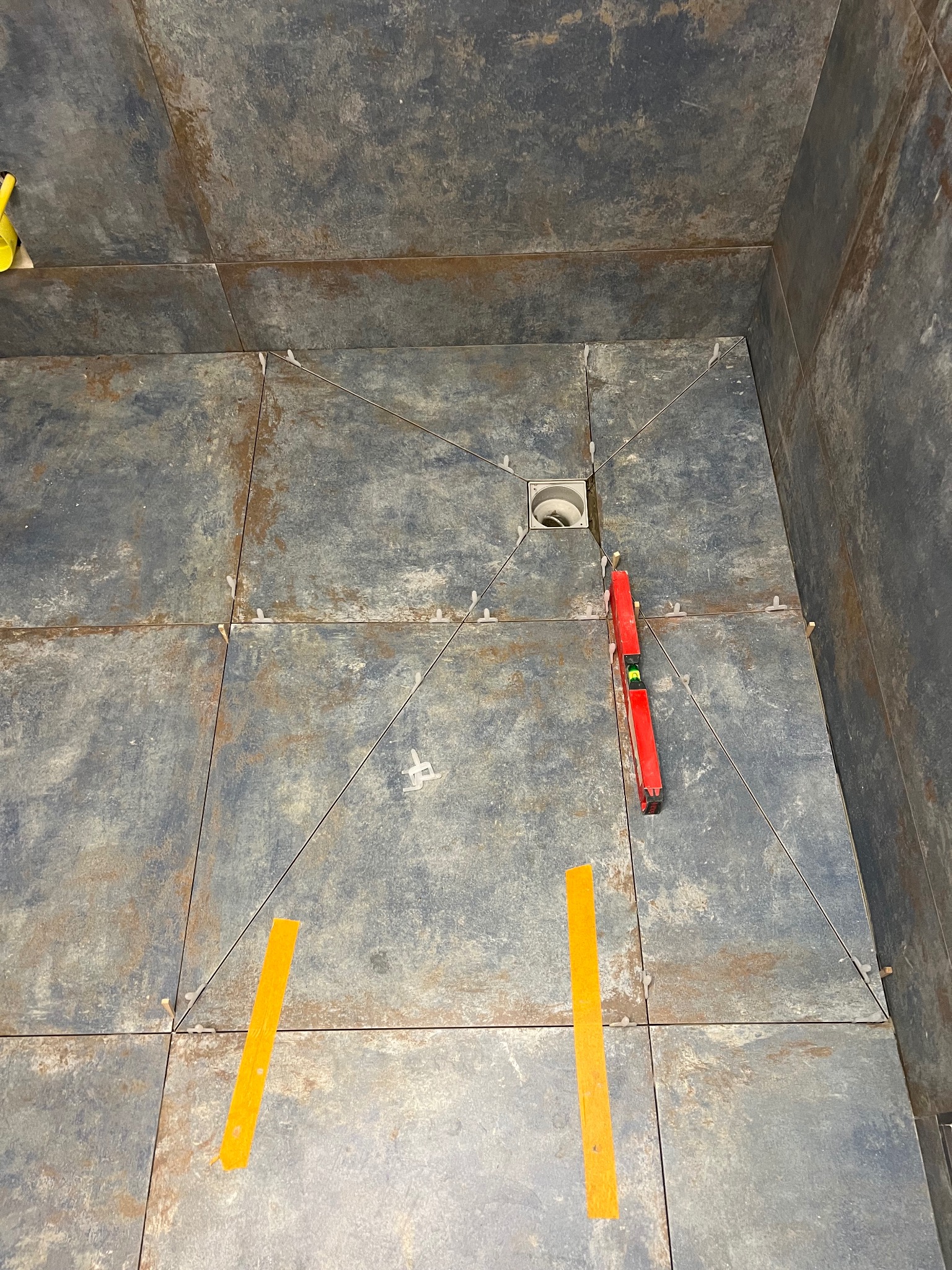 Ułożone, szare płytki w kabinie prysznicowej z odpływem liniowym. Widoczne poziomica, taśma malarska i krzyżyki dystansowe. Płytki imitują metal.