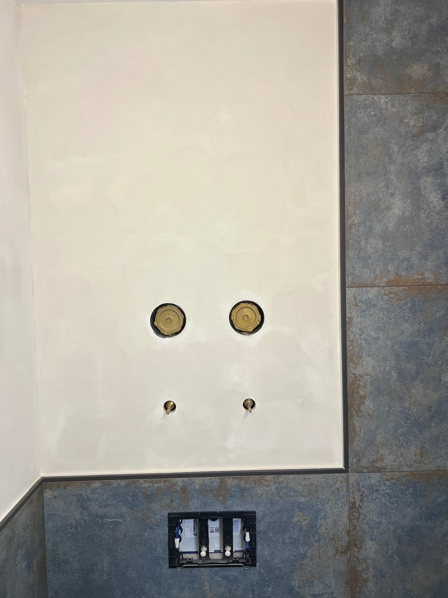 Ściana w łazience z przygotowanymi otworami na baterie i odpływ, oraz zabudowany stelaż podtynkowy WC. Widoczne płytki imitujące rdzę.