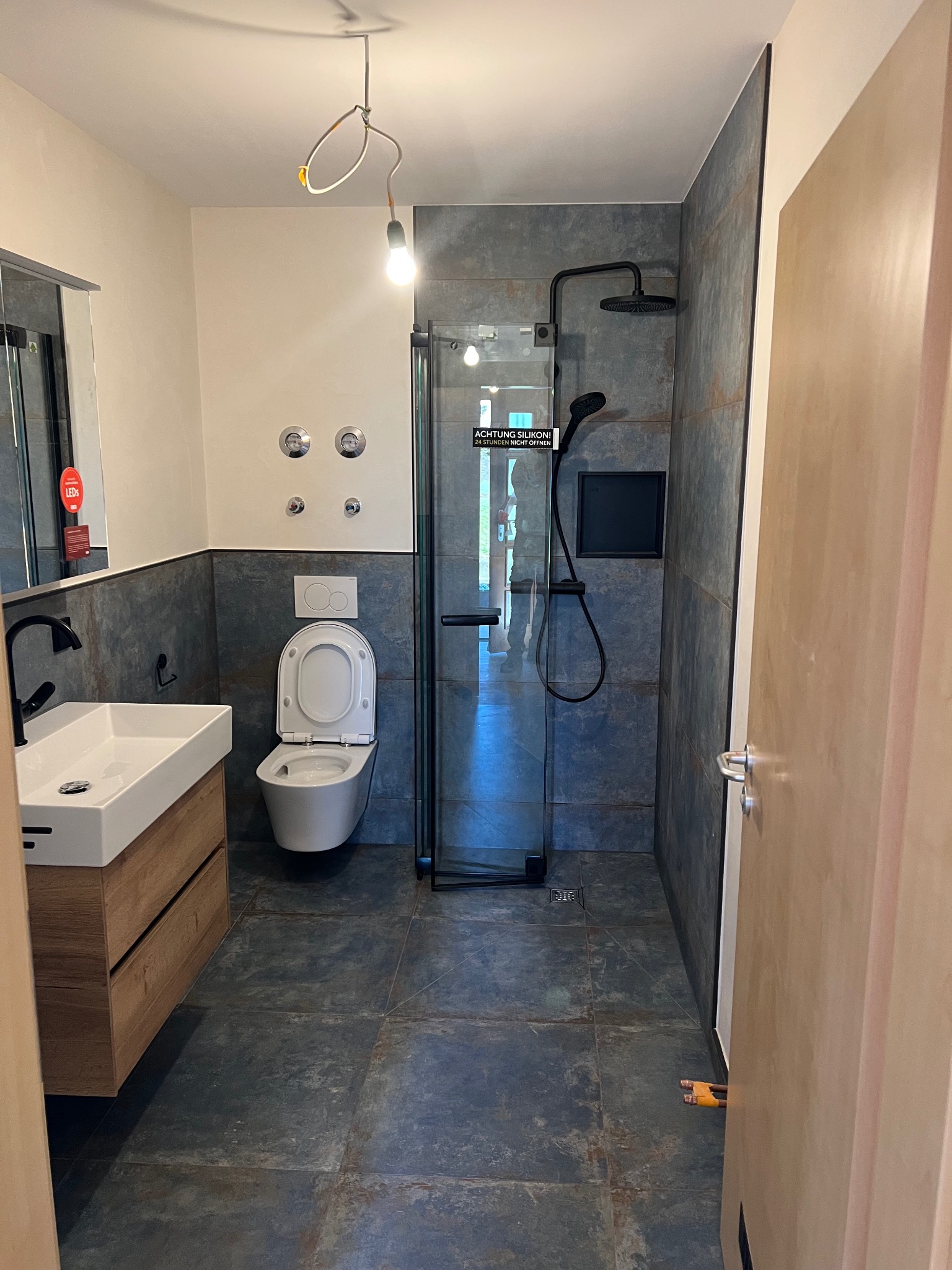 Nowoczesna łazienka z kabiną prysznicową typu walk-in, umywalką na drewnianej szafce i toaletą. Ciemne płytki imitujące kamień na ścianach i podłodze.