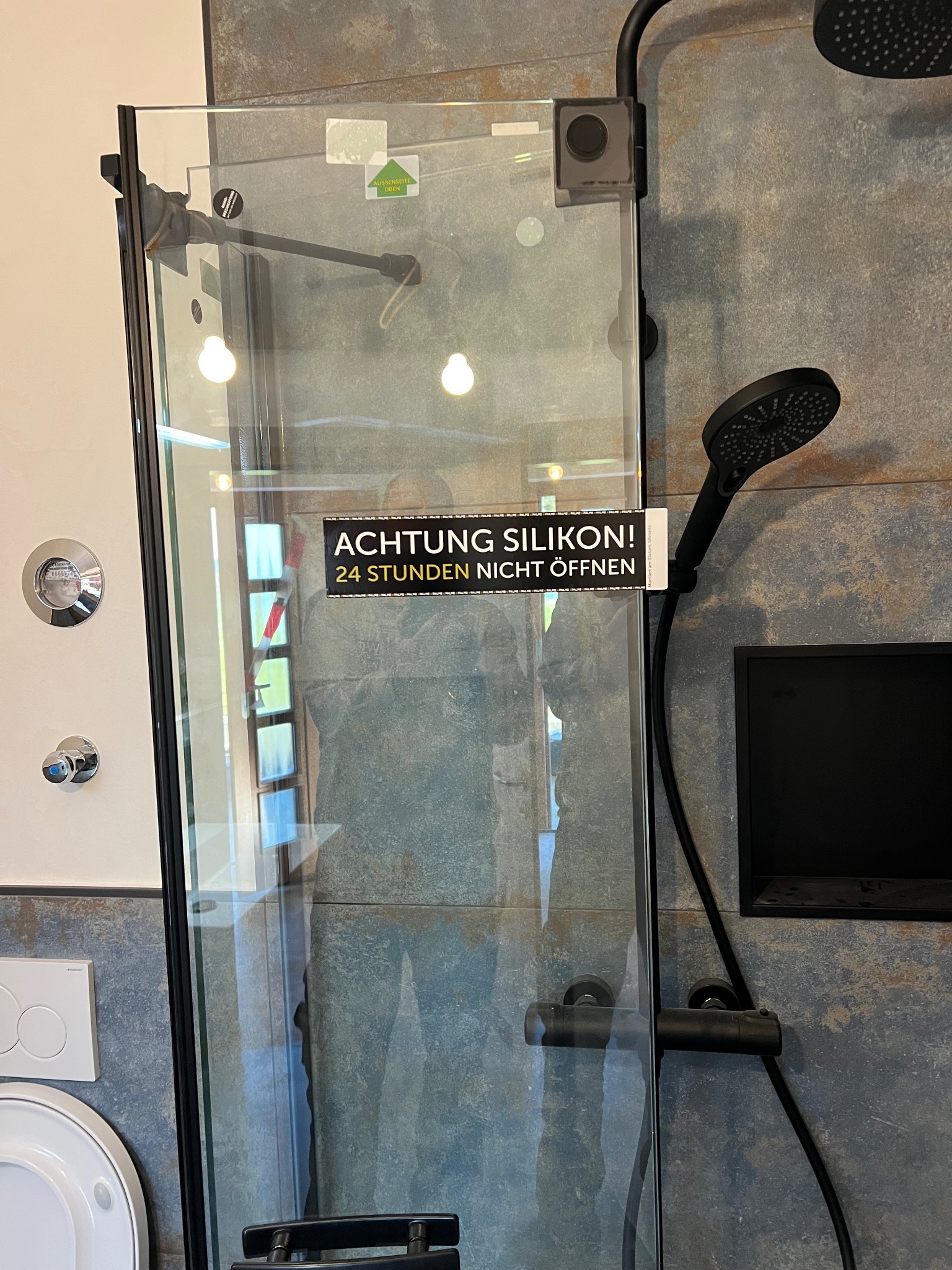 Nowoczesna kabina prysznicowa z czarnymi elementami, świeżo uszczelniona silikonem, o czym informuje naklejka. Widoczna armatura i płytki w odcieniach szarości.