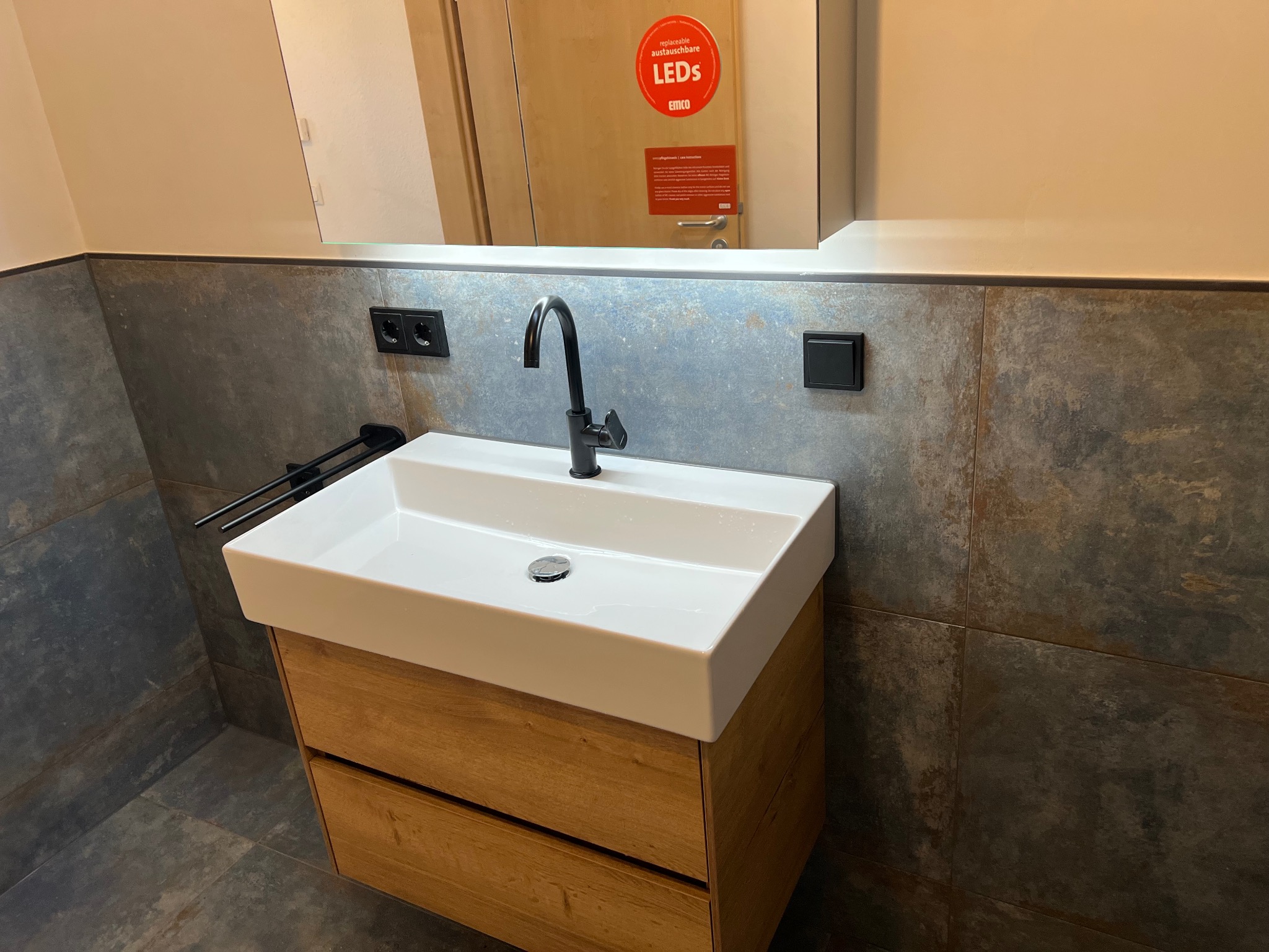 Nowoczesna łazienka: biała umywalka nablatowa na drewnianej szafce, czarna bateria, szare płytki imitujące kamień, czarne gniazdka i włączniki. Lustro z oświetleniem LED.