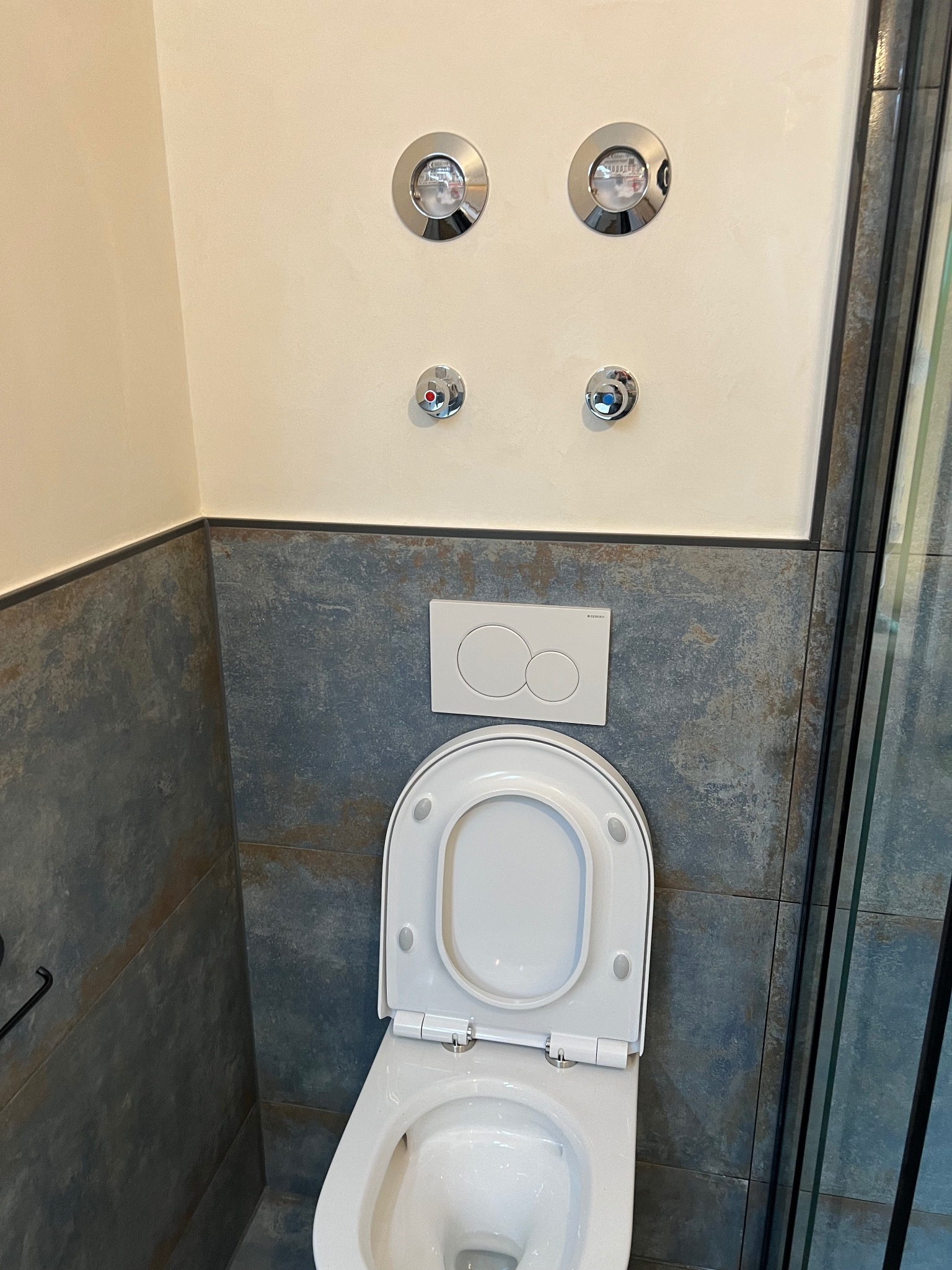 Nowoczesna toaleta z szarą, teksturowaną glazurą, chromowanymi przyciskami i armaturą. Widoczny fragment kabiny prysznicowej z czarną ramą. Minimalistyczny design.