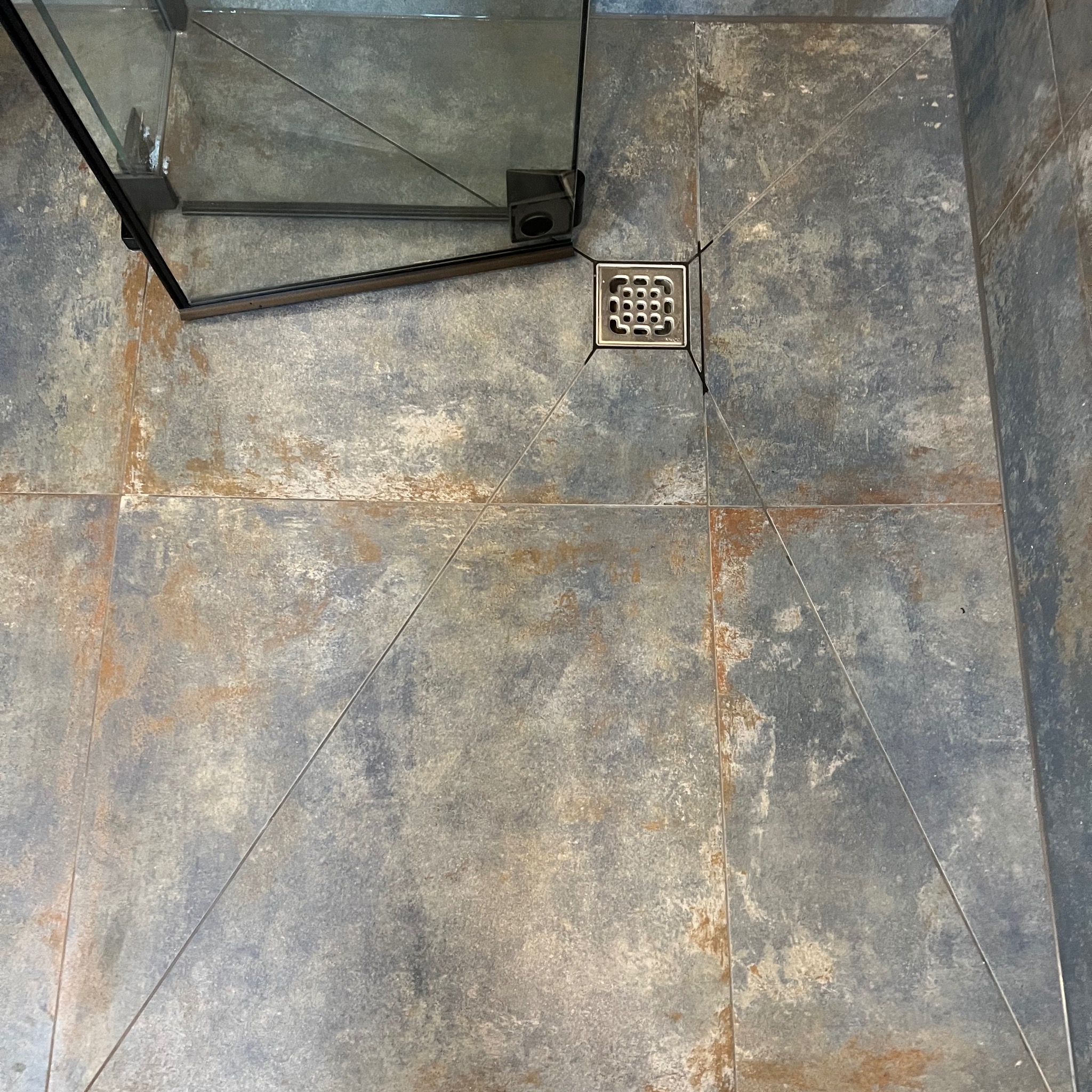 Szare płytki podłogowe w kabinie prysznicowej z odpływem liniowym. Widoczna część szklanej kabiny. Płytki imitują beton z rdzawymi przebarwieniami.