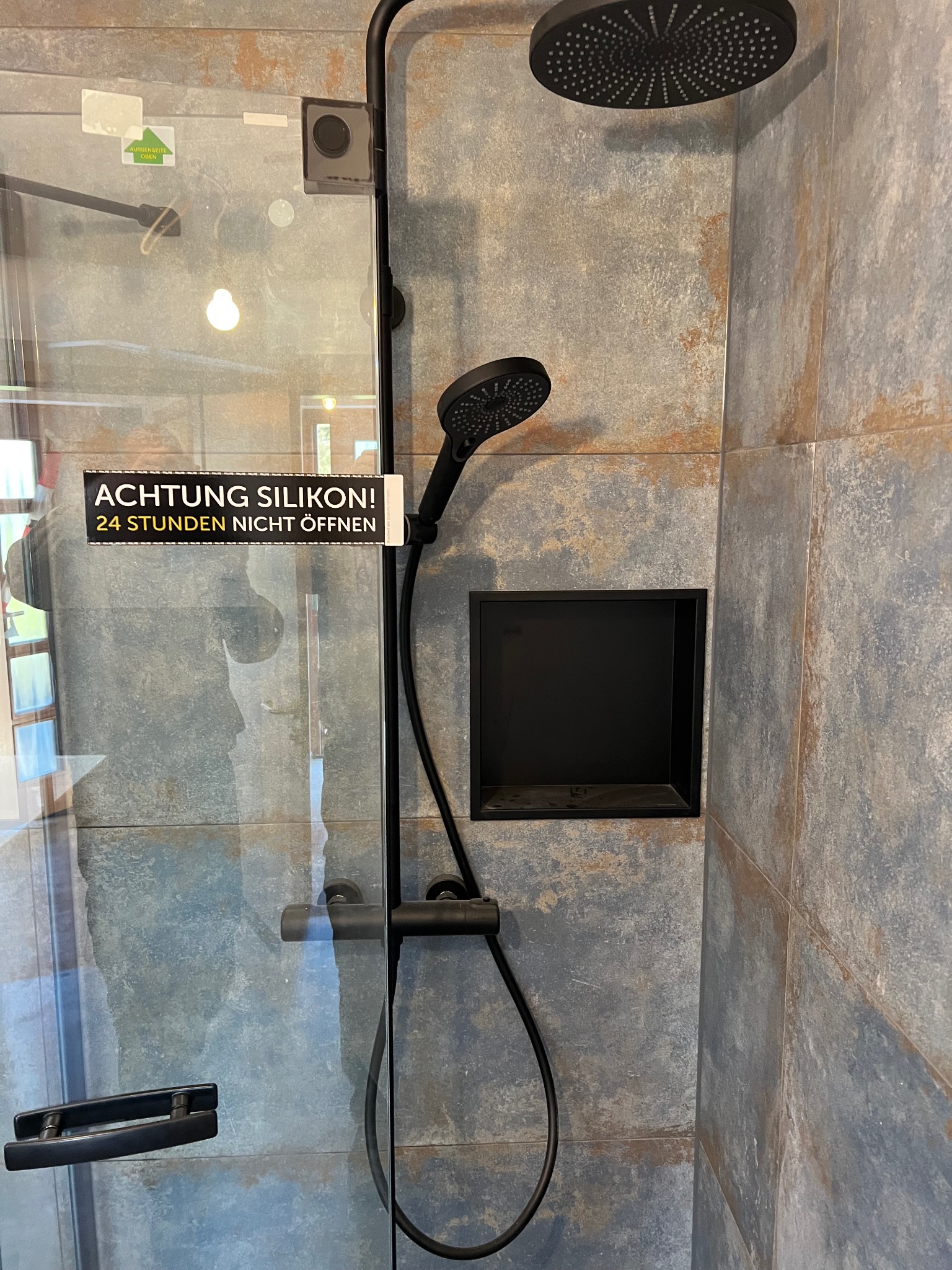 Nowoczesna kabina prysznicowa z czarną armaturą i wnęką w ścianie wyłożonej płytkami imitującymi kamień. Na szklanej ściance naklejka informująca o świeżym silikonie.