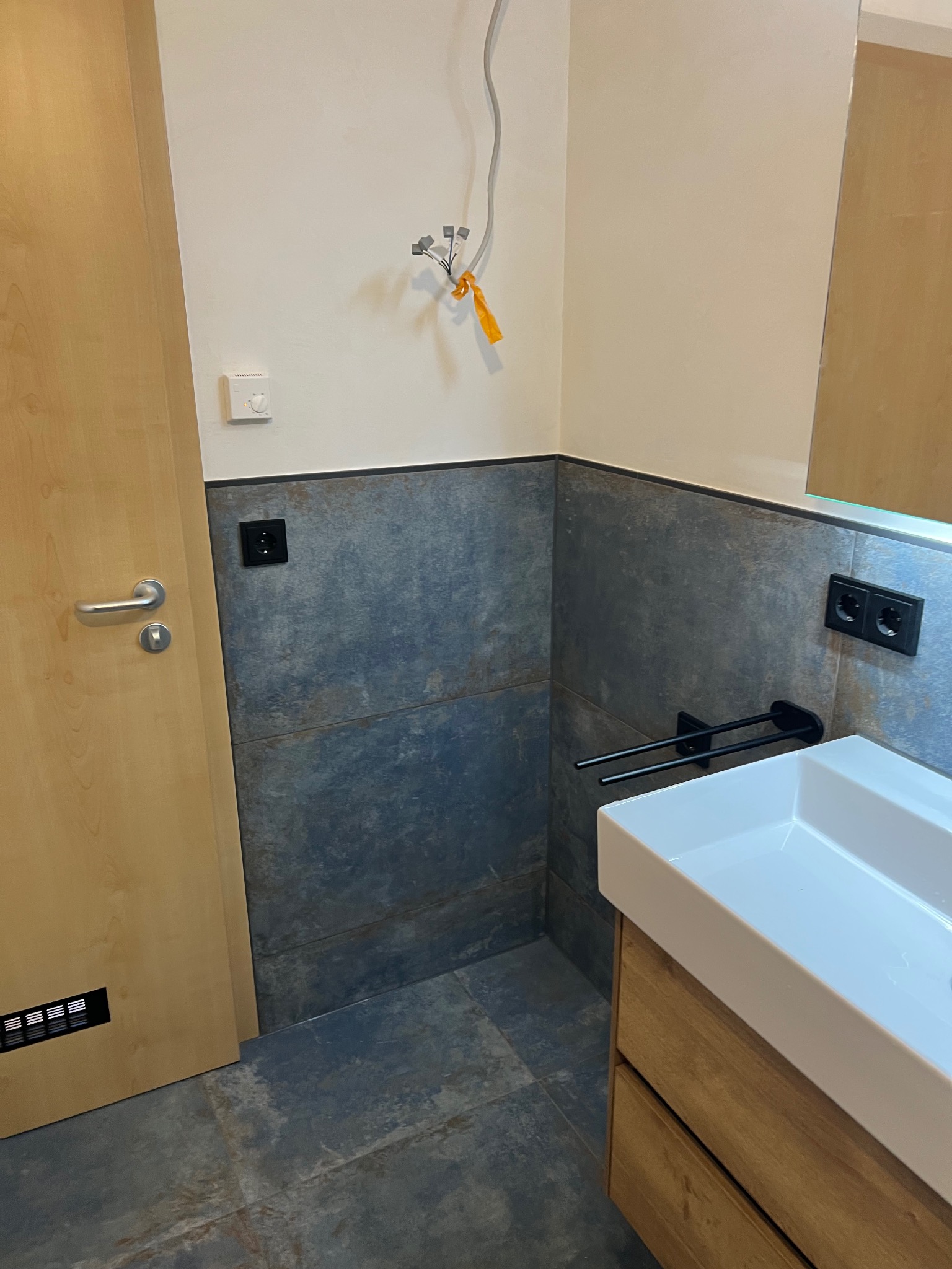 Nowoczesna łazienka z umywalką nablatową, szarą płytką imitującą kamień na ścianach i podłodze. Widoczne gniazdka elektryczne i fragment instalacji.