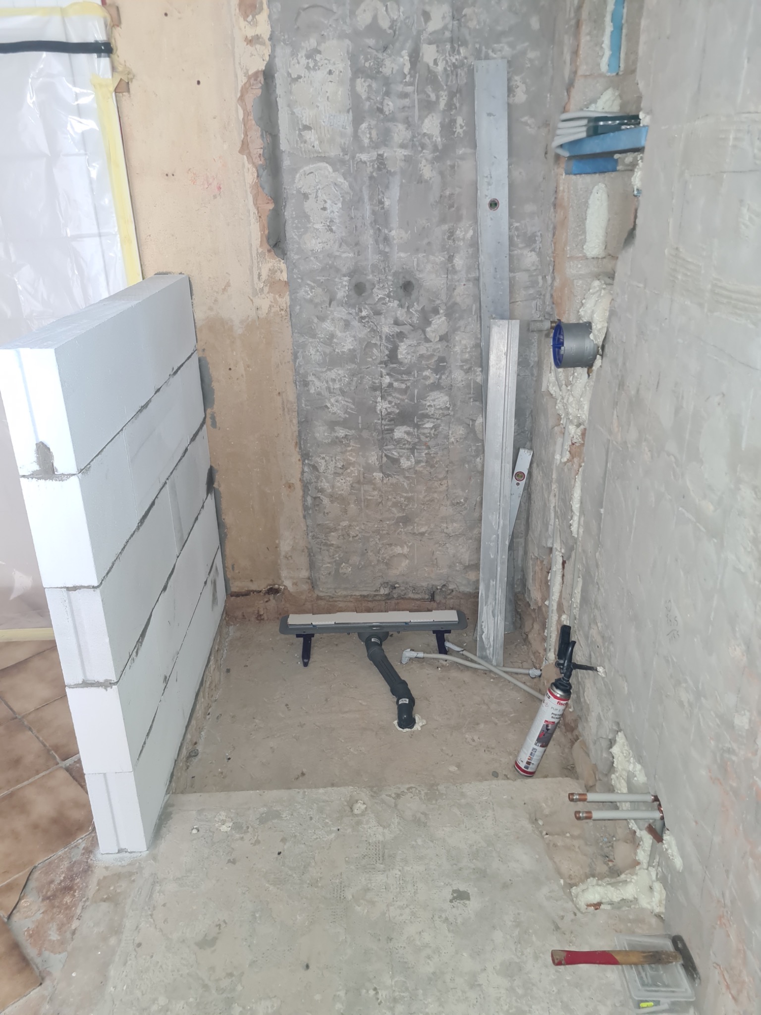 Wnętrze łazienki w trakcie remontu: widoczne bloczki Ytong, odpływ liniowy, poziomica, puszka pod baterię, pianka montażowa i fragment instalacji wodnej.