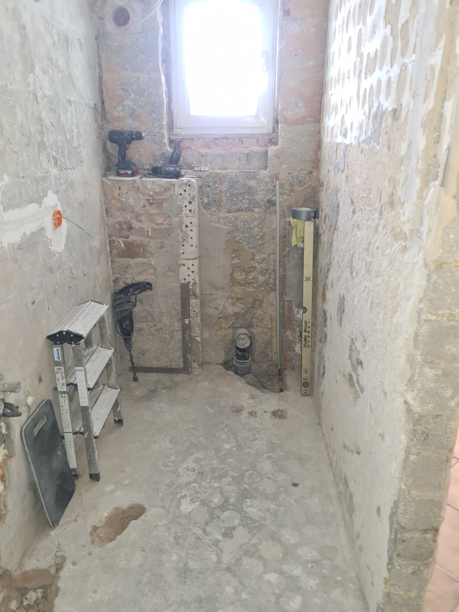 Wnętrze łazienki w trakcie demontażu: odsłonięte cegły, resztki tynku, narzędzia (wiertarki, poziomica), drabina. Widoczne instalacje. Okno w tle.