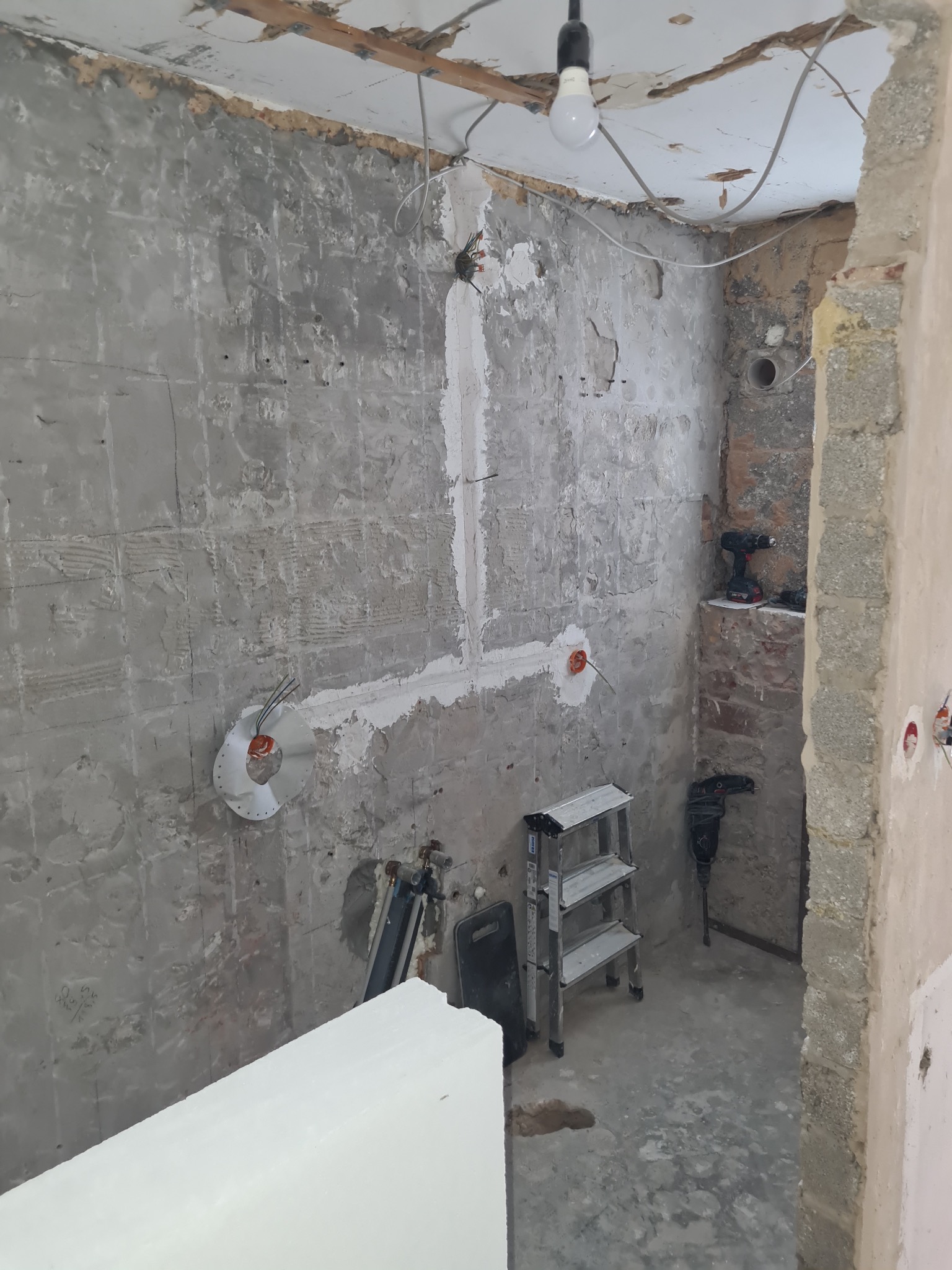 Remont łazienki w trakcie: ściany z odkrytą instalacją elektryczną i hydrauliczną, drabina, narzędzia. Widoczne ślady po skuciu płytek i przygotowaniu podłoża.