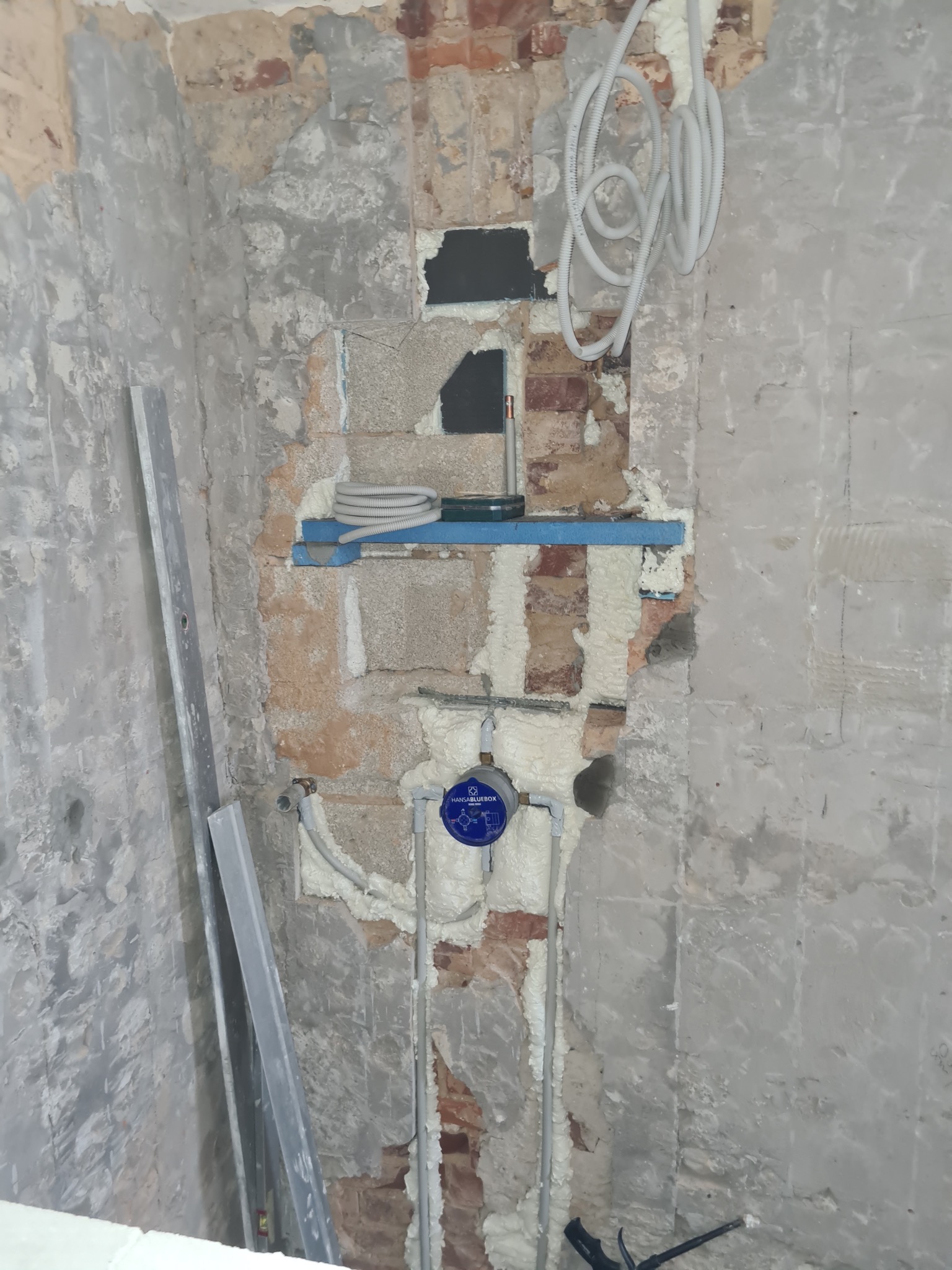 Ściana w trakcie remontu z widocznymi instalacjami wodno-kanalizacyjnymi, zabezpieczonymi pianką montażową i elementami konstrukcyjnymi. Widoczne narzędzia i materiały budowlane.