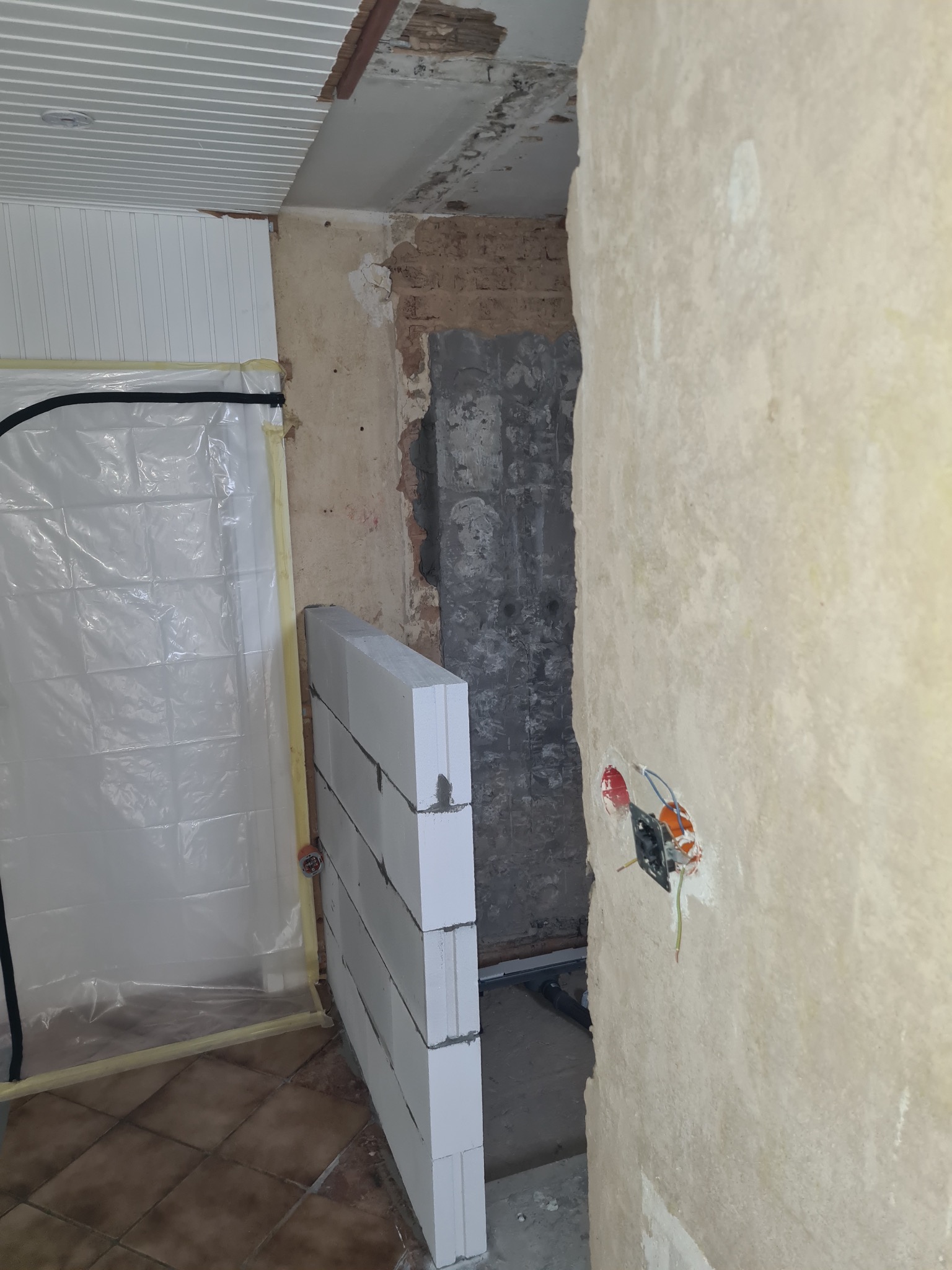 Wnętrze w trakcie prac: ściana z pustaków, zabezpieczona folią strefa, widoczne przewody elektryczne. Fragment sufitu z boazerią. Etap budowy lub remontu.