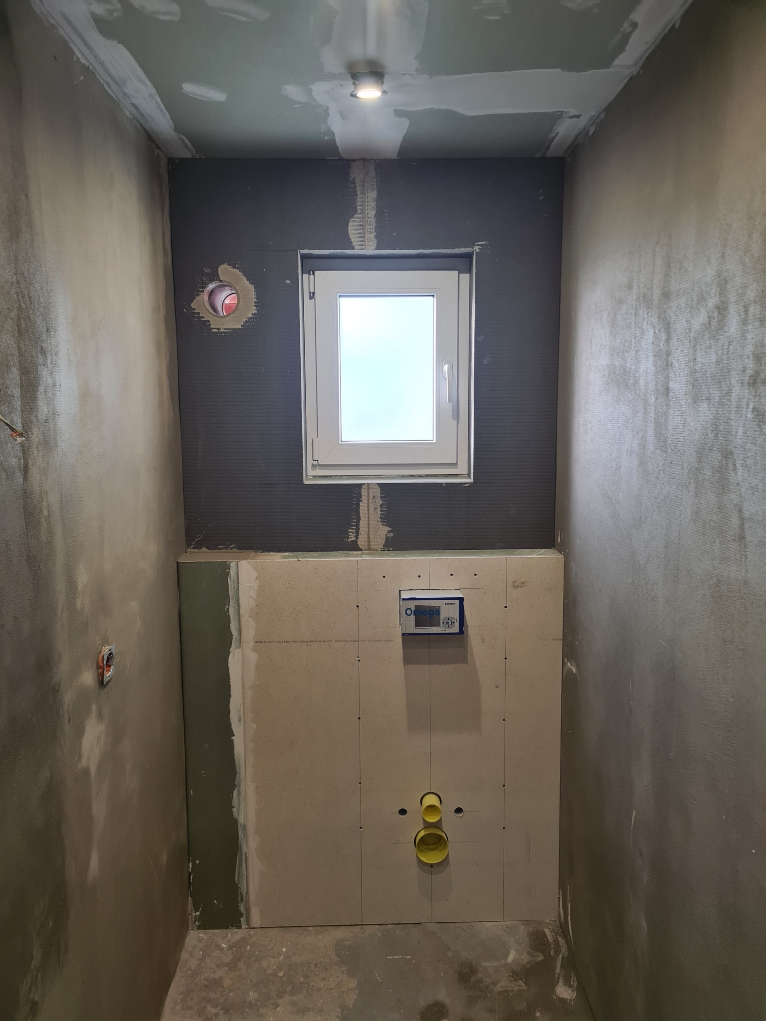 Łazienka w trakcie remontu: ściany z surowym tynkiem, okno, zabudowa stelaża WC z płyt karton-gips, widoczne instalacje. Etap prac wykończeniowych.