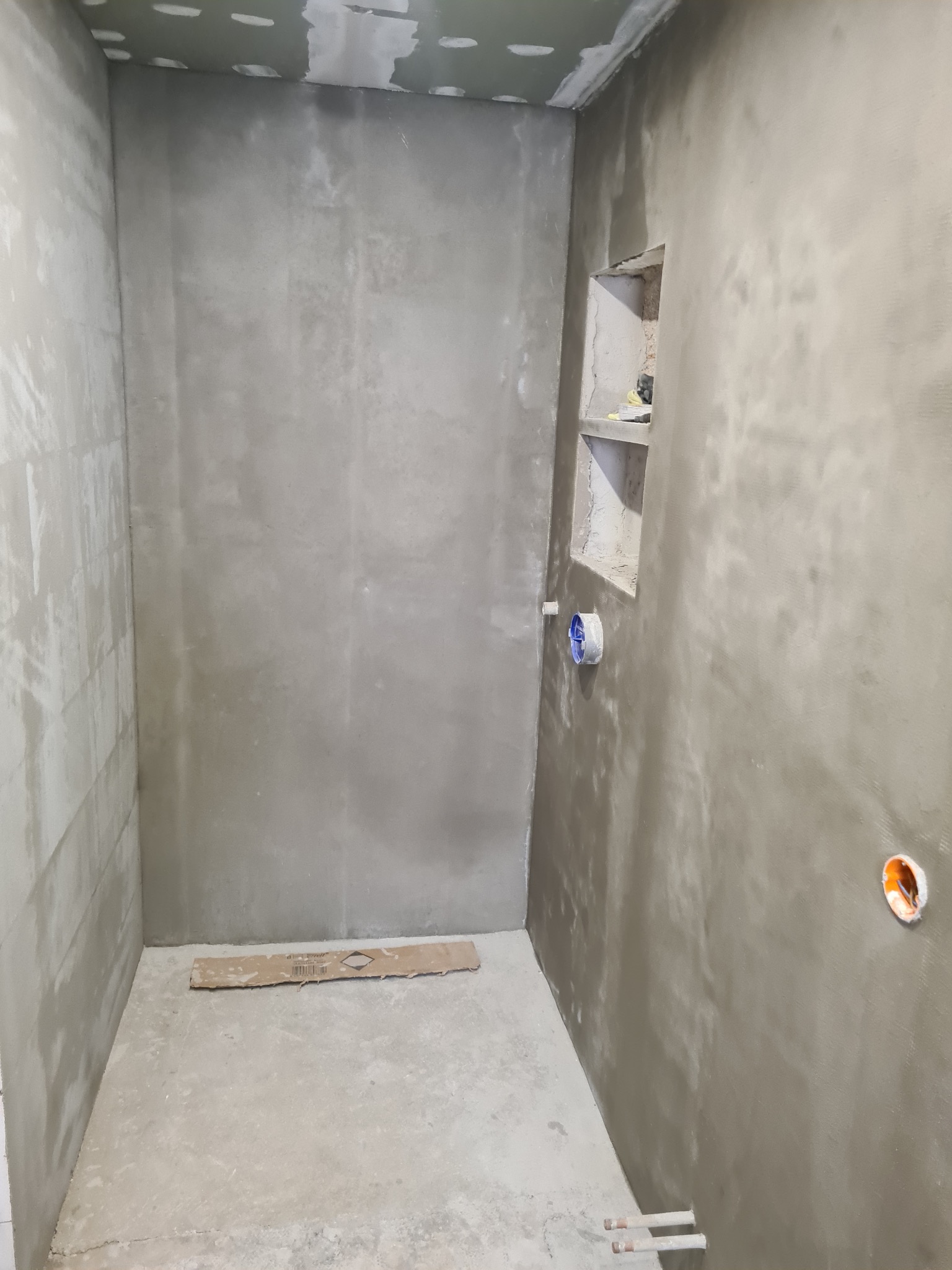 Surowa łazienka w trakcie prac wykończeniowych: otynkowane ściany, przygotowane instalacje, wnęka w ścianie. Widoczne ślady po łatach na suficie.