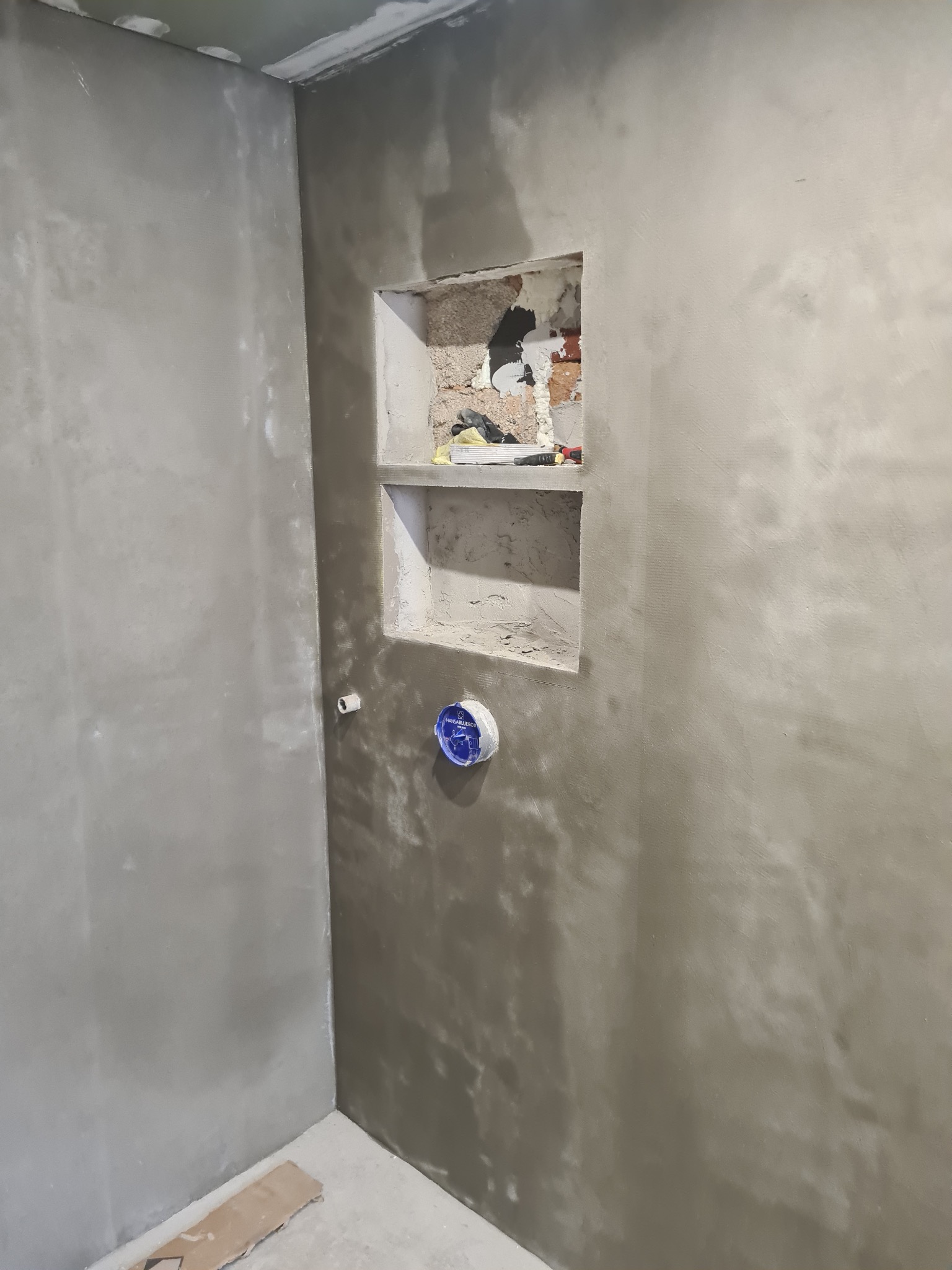 Ściana w trakcie szpachlowania z wnęką i puszką elektryczną. Widoczne ślady szpachli i niedoskonałości, co wskazuje na etap prac wykończeniowych w pomieszczeniu.