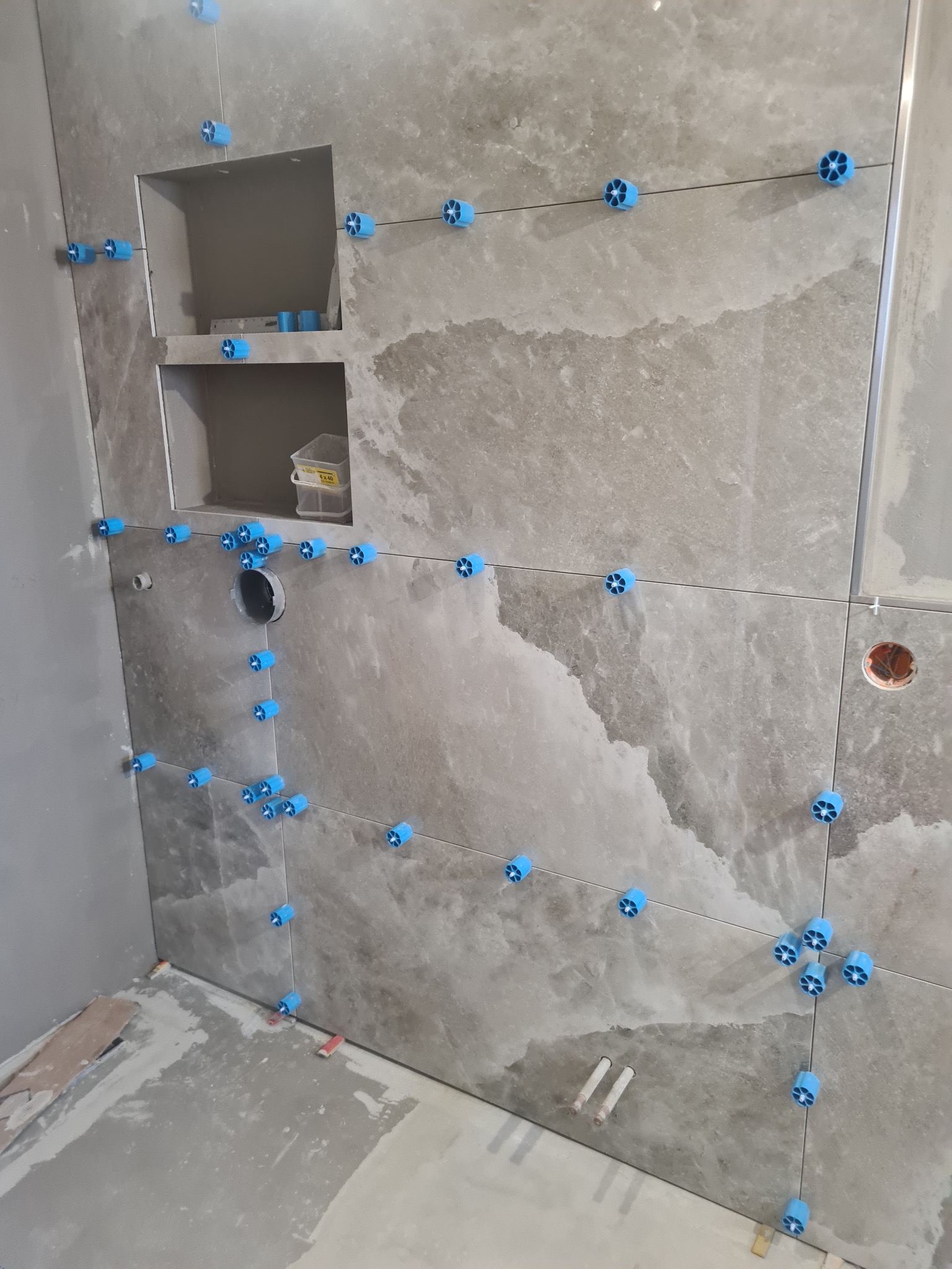 Ściana w trakcie układania płytek w łazience. Widoczne niebieskie klipsy poziomujące i wnęki w ścianie. Szare płytki imitujące kamień.