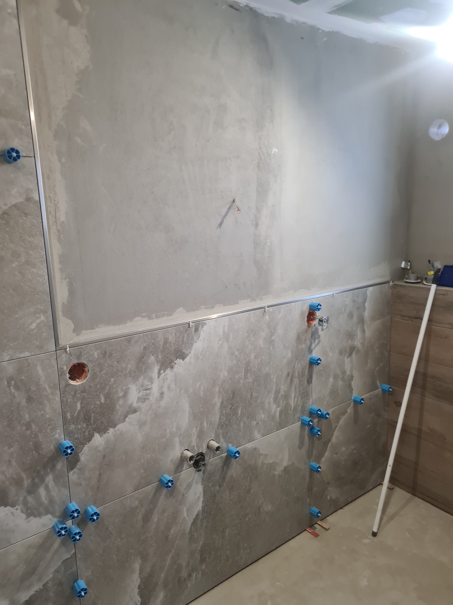 Układanie płytek w łazience z użyciem systemu poziomowania. Widoczne płytki imitujące marmur, listwa dekoracyjna i przygotowane punkty instalacyjne.