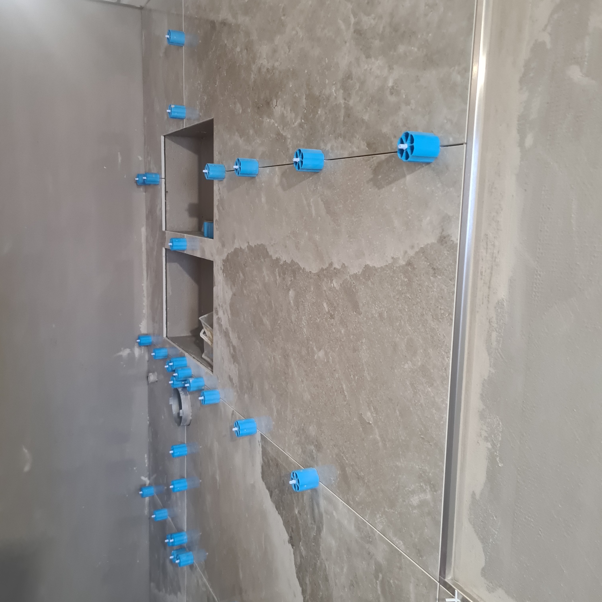 Ściana łazienki w trakcie wykańczania z szarymi płytkami imitującymi kamień, poziomowana za pomocą niebieskich klipsów i sznurka. Widoczne wnęki w ścianie.