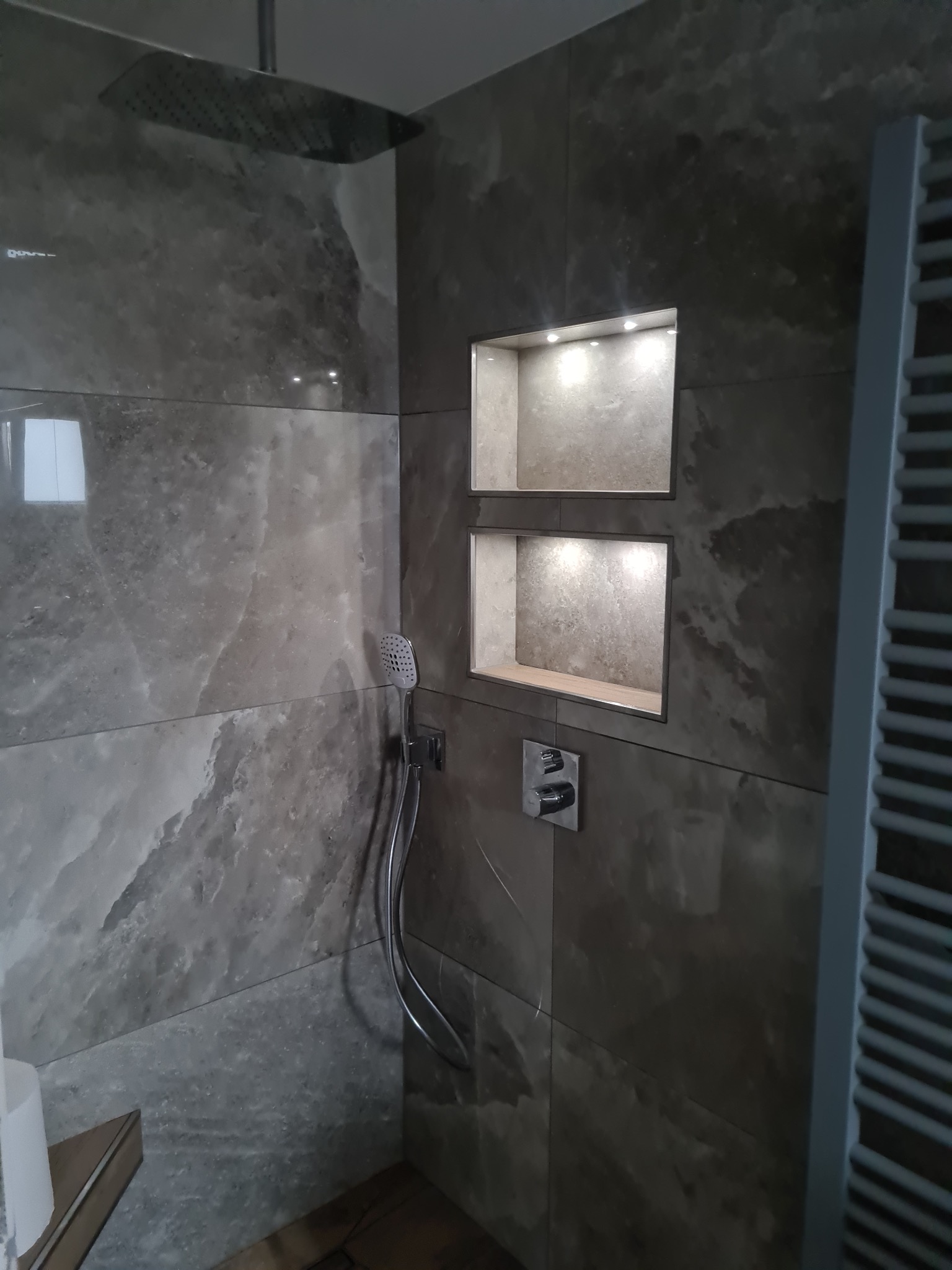 Nowoczesna kabina prysznicowa z szarymi płytkami imitującymi kamień, wnękami z oświetleniem LED i deszczownicą. Minimalistyczny design z grzejnikiem drabinkowym.