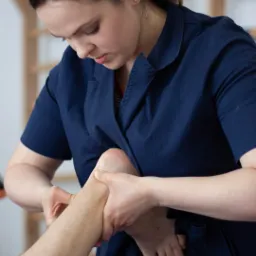 Kobieta w granatowej bluzie uciska stopę pacjenta podczas sesji w gabinecie w Kościerzynie, drabinka gimnastyczna w tle.