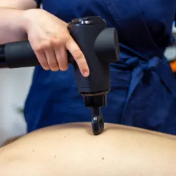 Osoba w niebieskim uniformie używa czarnego pistoletu do masażu na plecach pacjenta, widoczne dłonie i fragment sylwetki pacjenta.