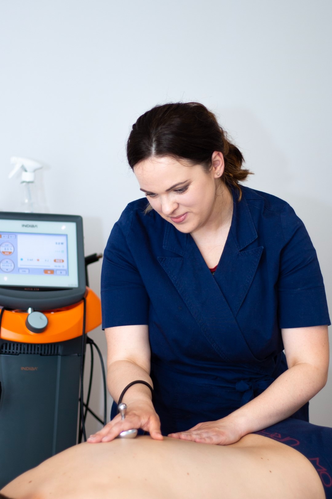 Kobieta w granatowym uniformie przeprowadza zabieg na plecach pacjenta za pomocą urządzenia Indiba Activ CT9, z widocznym ekranem dotykowym i aplikatorem.