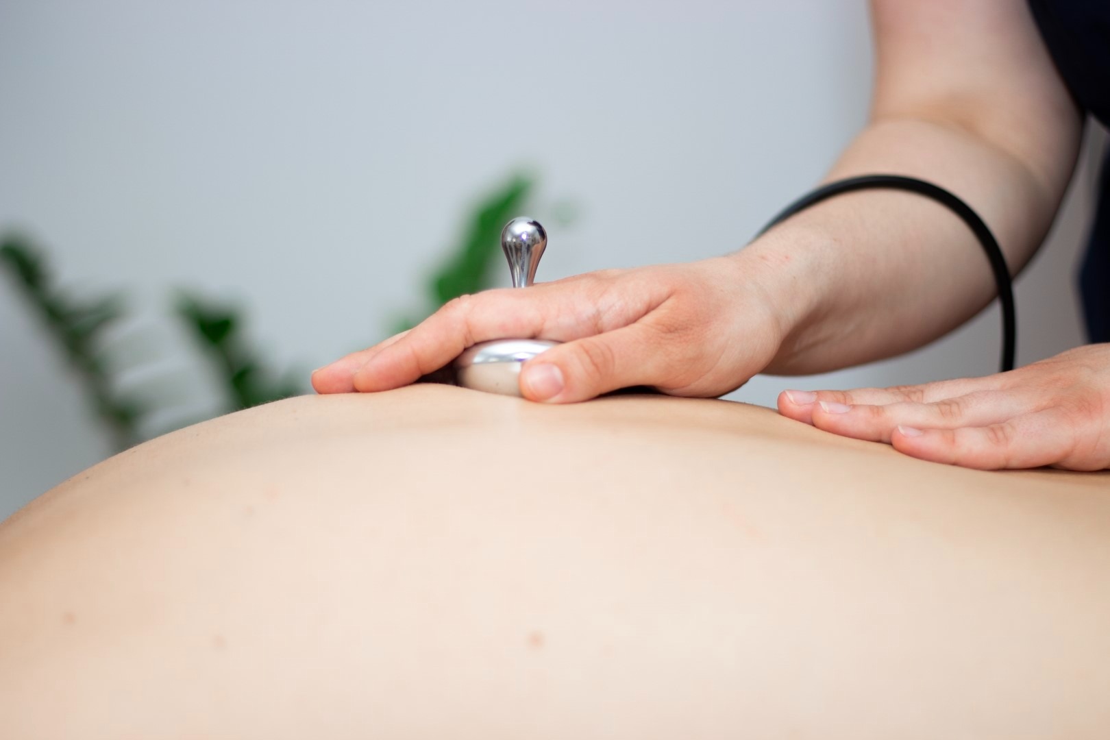Terapia dźwiękiem: metalowy aplikator z uchwytem przykładany do pleców pacjenta, dłoń terapeuty stabilizuje urządzenie, widoczne tło z rośliną.