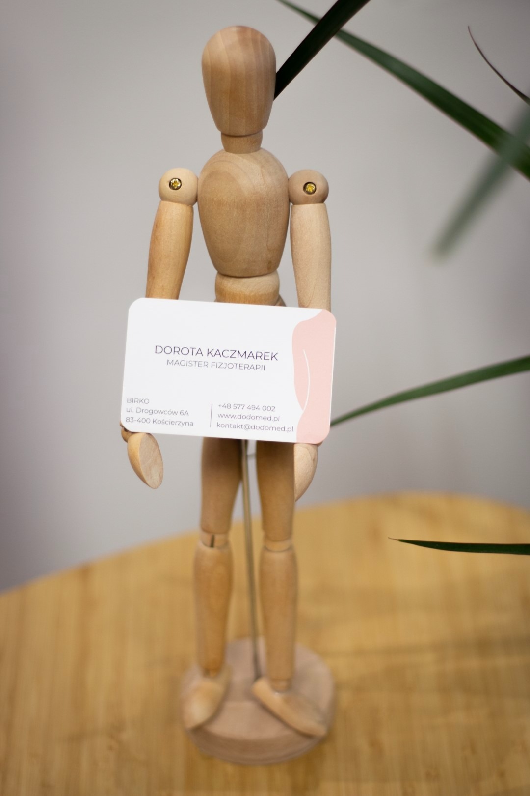 Drewniany model człowieka trzymający wizytówkę z danymi kontaktowymi magistra fizjoterapii Doroty Kaczmarek, na jasnym tle z elementami roślinnymi.