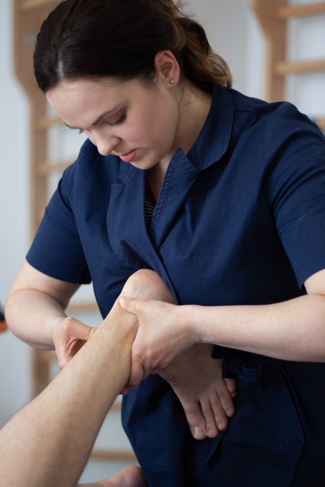 Kobieta w granatowej bluzie uciska stopę pacjenta podczas sesji w gabinecie w Kościerzynie, drabinka gimnastyczna w tle.