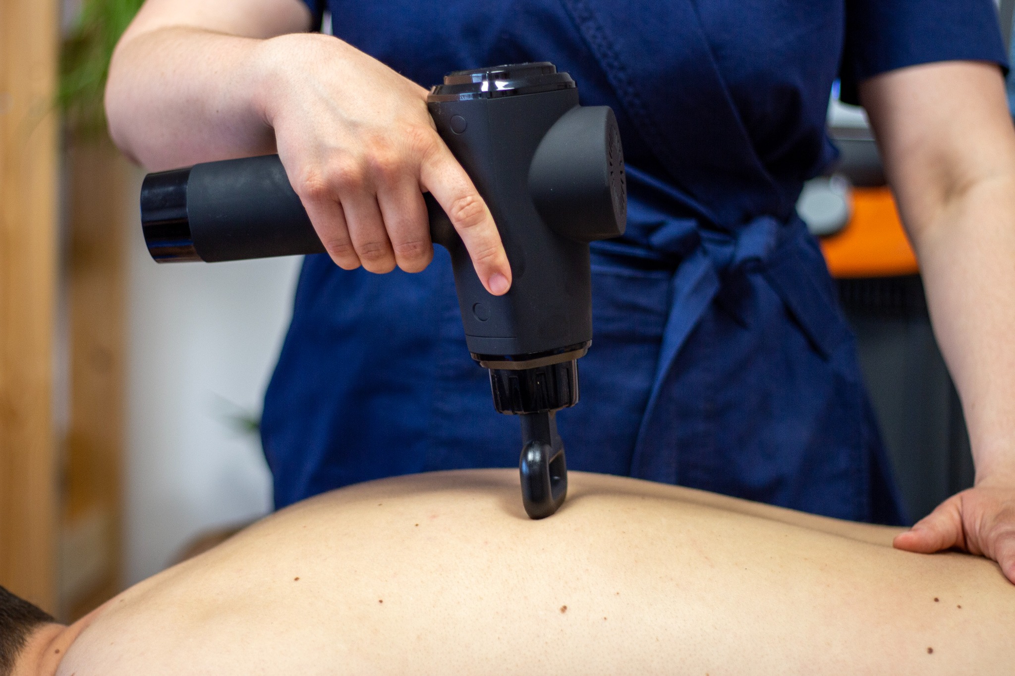 Osoba w niebieskim uniformie używa czarnego pistoletu do masażu na plecach pacjenta, widoczne dłonie i fragment sylwetki pacjenta.