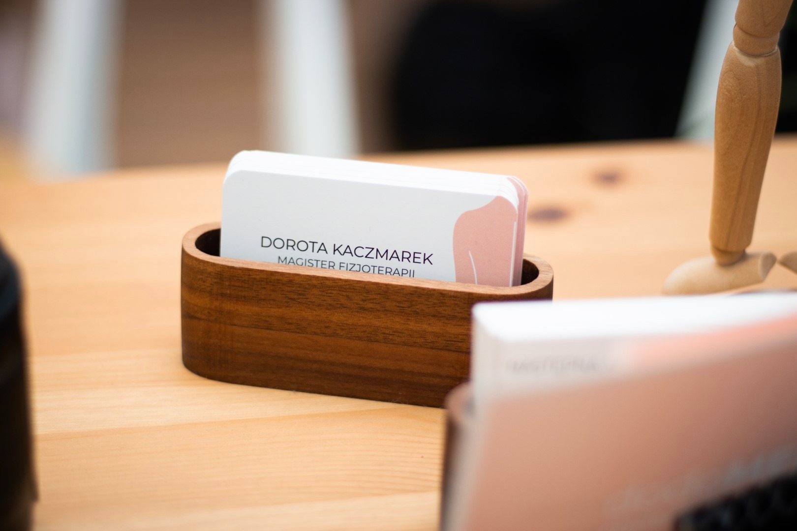 Wizytówki Doroty Kaczmarek, magister fizjoterapii, umieszczone w drewnianym pojemniku, z delikatnym rysunkiem pleców w kolorze pudrowego różu, obok drewnianego modelu anatomicznego.