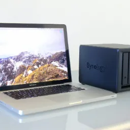 Laptop MacBook Pro podłączony kablem do serwera NAS Synology DS418play na błyszczącym stole, z widocznym ekranem laptopa wyświetlającym górski krajobraz.