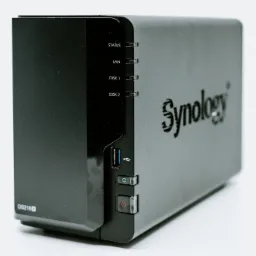 Czarna obudowa serwera NAS Synology DS218+ z widocznymi diodami statusu, portem USB i przyciskiem zasilania na białym tle.
