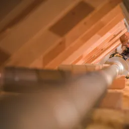 Instalator w okularach ochronnych i rękawicach montuje rurę wentylacyjną w przestrzeni pod dachem, widoczne drewniane elementy konstrukcyjne dachu i izolacja.