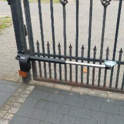 Częściowo otwarta, czarna, metalowa brama ze zdobieniami w postaci spiral i ostrych zakończeń, wyposażona w siłownik FAAC, widoczny na tle szarej kostki brukowej i ulicy.