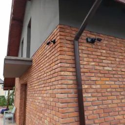 Zewnętrzna ściana budynku z czerwonej cegły klinkierowej, z zamontowanymi dwoma czarnymi kamerami monitoringu i brązową rynną spustową. Widoczne szare elementy elewacji i drewniane wykończenie dachu.