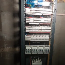 Rozdzielnica elektryczna w metalowej szafie, widoczne liczne zabezpieczenia nadprądowe, różnicowoprądowe i listwy zaciskowe, instalacja w surowym stanie.