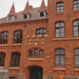 Odnowiona elewacja budynku z czerwonej cegły z charakterystycznymi zdobieniami i nową stolarką okienną, zabezpieczone wejście do budynku folią.