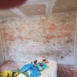 Częściowo otynkowana ściana z cegły w trakcie remontu, z widocznymi śladami białej farby i workami z folii budowlanej na podłodze.