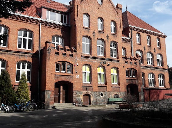 Fasada budynku z czerwonej cegły z licznymi oknami, widoczne rowery zaparkowane przed budynkiem.