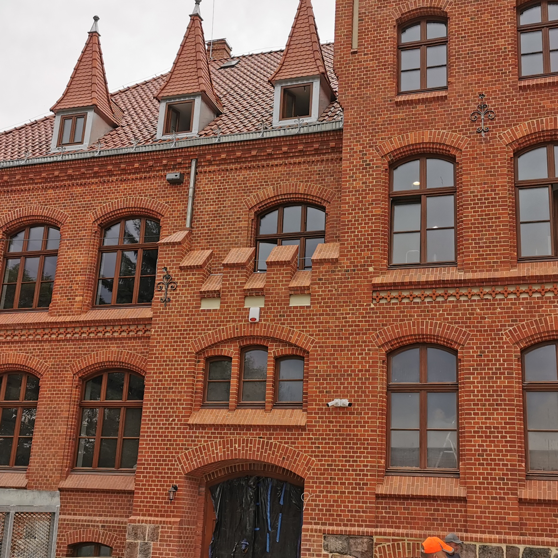 Odnowiona elewacja budynku z czerwonej cegły z charakterystycznymi zdobieniami i nową stolarką okienną, zabezpieczone wejście do budynku folią.