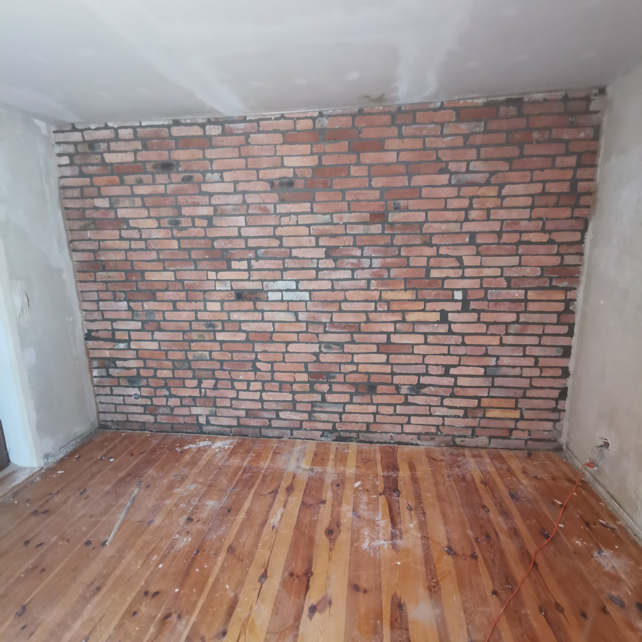 Ściana z czerwonej cegły w surowym wnętrzu, widoczne ślady po pracach remontowych na podłodze i ścianach, gniazdko elektryczne wystające ze ściany.