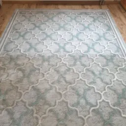 Prostokątny dywan z marokańskim wzorem w odcieniach szarości i błękitu na drewnianej podłodze, widoczny grzejnik ścienny w tle.